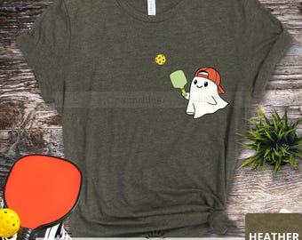 May include: A green t-shirt with a ghost wearing a red hat and holding a green paddle ball racquet. The ghost is playing paddle ball with a yellow ball. The text "sunshine pocket" is printed on the bottom of the shirt. The shirt is heather olive.