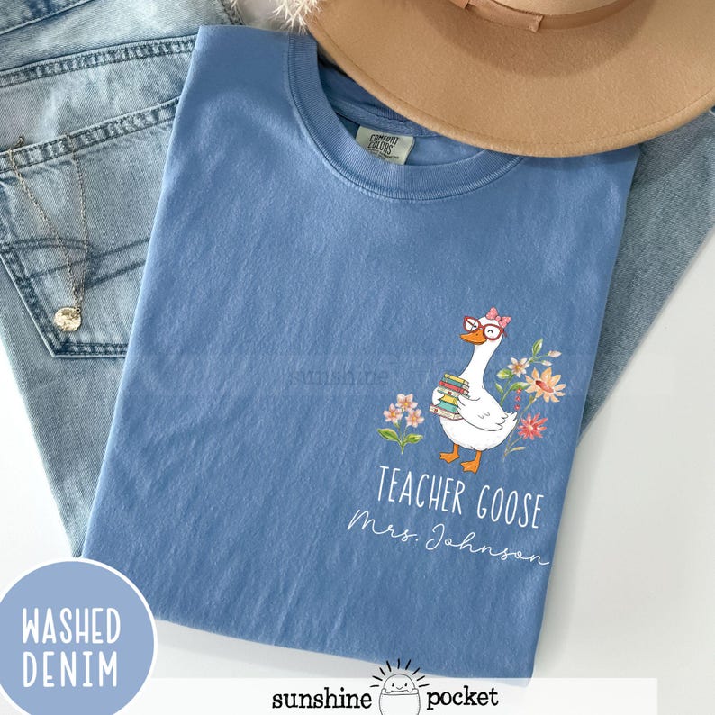 May include: A pale blue t-shirt with a cartoon goose wearing glasses and holding books, surrounded by flowers. The text "Teacher Goose Mrs. Johnson" is printed below the goose. The shirt is folded on top of denim jeans.