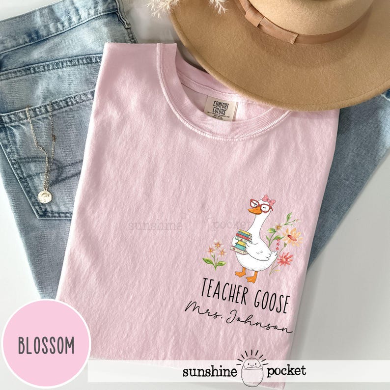 May include: A light pink t-shirt featuring a goose wearing glasses and holding books, with the text "Teacher Goose Mrs. Johnson". The shirt is part of the "Blossom" collection.
