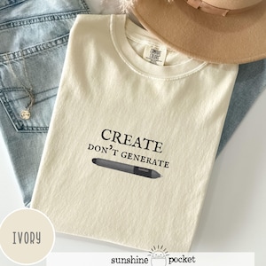 May include: Ivory-colored t-shirt with the text "CREATE DON'T GENERATE" and a graphic of a digital pen. The shirt is a Comfort Colors brand. The shirt is displayed on a light blue denim background with a necklace and a tan hat.