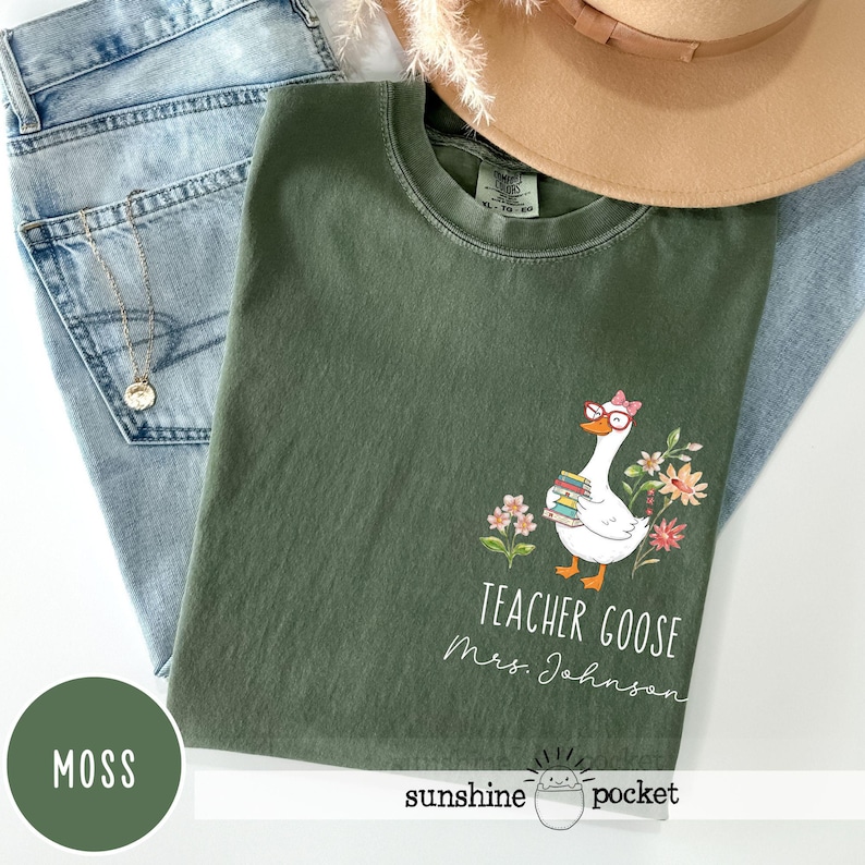 May include: A moss green t-shirt featuring a goose graphic wearing glasses and carrying books, with flowers. The text reads "Teacher Goose Mrs. Johnson". The shirt is folded on top of blue jeans. A hat is visible.