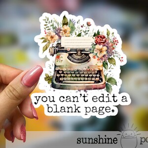 May include: A sticker featuring a vintage typewriter with floral accents. The typewriter is cream-colored with black keys and is surrounded by flowers in shades of pink, yellow, and purple. The text "you can't edit a blank page" is below.