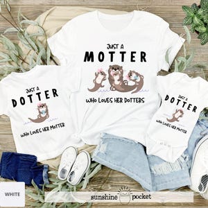 May include: White t-shirts and a onesie with otter graphics. The shirts have the text "JUST A MOTTER WHO LOVES HER DOTTERS" and "JUST A DOTTER WHO LOVES HER MOTTER". The onesie also has the text "JUST A DOTTER WHO LOVES HER MOTTER".
