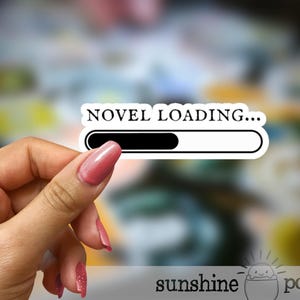 May include: A white sticker with the text "NOVEL LOADING..." and a progress bar, held between fingers with pink nail polish. The sticker has a black progress bar that is partially filled. The background is blurred with various colors.