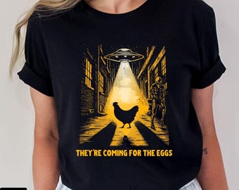 May include: Black t-shirt with a humorous graphic. The design features a chicken illuminated by a UFO beam in an alley, with an alien and the text "THEY'RE COMING FOR THE EGGS".