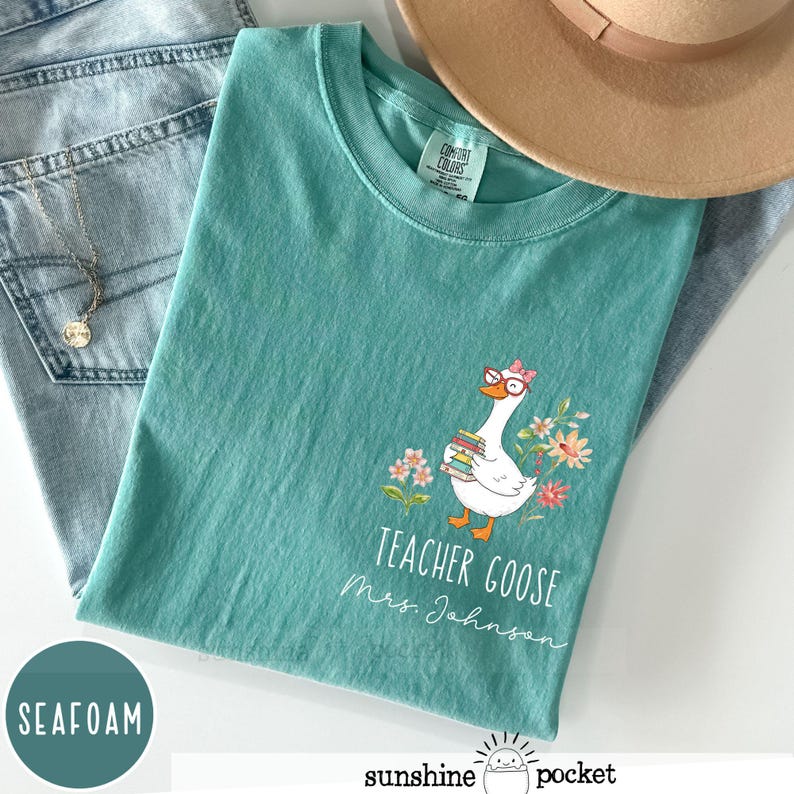 May include: Seafoam green t-shirt featuring a goose wearing glasses and carrying books, with floral accents. The text reads "Teacher Goose Mrs. Johnson". A tan hat and blue jeans are visible in the background. The t-shirt is made of a soft material.