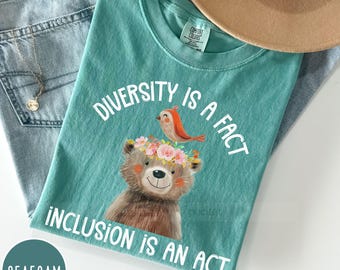 May include: Seafoam green t-shirt with a brown bear and a red bird graphic. The text "Diversity is a fact, Inclusion is an act" is printed on the shirt.