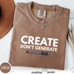 May include: Espresso-colored t-shirt with the text "CREATE DON'T GENERATE" in white, along with a graphic of a digital pen. The shirt has a Comfort Colors tag and a silver necklace. A brown hat is visible in the background.