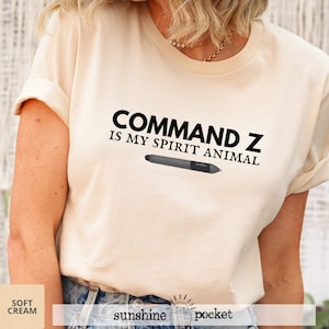 May include: Cream-colored t-shirt with the text "COMMAND Z IS MY SPIRIT ANIMAL" in black, along with a graphic of a digital pen. The shirt has short sleeves and a crew neck. The words "soft cream" and "sunshine pocket" are also printed on the shirt.