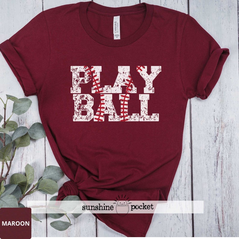 May include: Maroon t-shirt with a white distressed graphic that says "PLAY BALL" with a baseball graphic in the middle of the word "PLAY".
