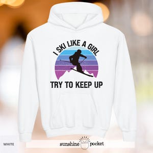 Peut inclure: Un sweat à capuche blanc avec l'inscription "I SKI LIKE A GIRL TRY TO KEEP UP". Le motif représente un skieur en silhouette devant un paysage de montagne violet et bleu et un soleil couchant.