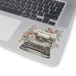 K&ouml;nnte beinhalten: Eine Vintage-Schreibmaschine mit floralen Akzenten und dem Zitat "Um zu beginnen, beginne." von William Wordsworth. Die Schreibmaschine ist blau und grau mit einer schwarzen Tastatur. Die Blumen sind rosa, wei&szlig; und gr&uuml;n.