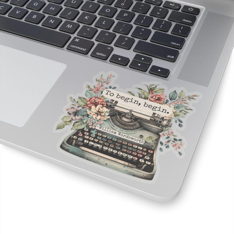K&ouml;nnte beinhalten: Eine Vintage-Schreibmaschine mit floralen Akzenten und dem Zitat "Um zu beginnen, beginne." von William Wordsworth. Die Schreibmaschine ist blau und grau mit einer schwarzen Tastatur. Die Blumen sind rosa, wei&szlig; und gr&uuml;n.