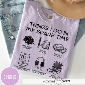 May include: Orchid-colored t-shirt with the text "THINGS I DO IN MY SPARE TIME" and illustrations of a book, pencil, headphones, and laptop. The shirt also has the text "sunshine pocket".