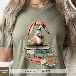 May include: Heather olive green t-shirt featuring a whimsical illustration. A bunny wearing glasses and a blue shirt sits atop books with the phrases "read books", "be kind", and "stay weird". A teacup with a heart is also present.