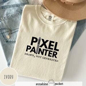 May include: Ivory-colored t-shirt with the words "PIXEL PAINTER" in bold black letters, with a graphic of a digital pen. Below the text is the phrase "DRAWN, NOT GENERATED". The shirt is folded on top of blue jeans.