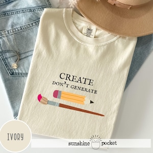 May include: An ivory t-shirt with a crew neckline featuring the black text "CREATE DON'T GENERATE" above a graphic of a yellow pencil and a paintbrush with a pink tip. The shirt has a Comfort Colors label at the inner neck and a short sleeve design.