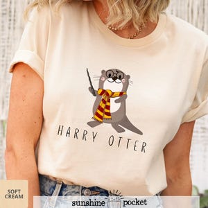 May include: A cream-colored t-shirt featuring a graphic of a brown otter wearing round glasses and a red and yellow striped scarf while holding a black wand. Below the otter, the text HARRY OTTER is printed in a simple black sans-serif font.