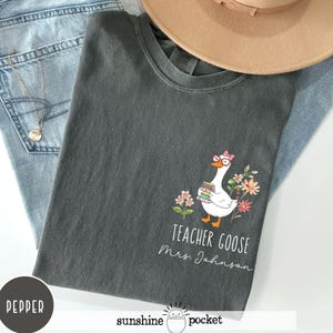 May include: Dark grey t-shirt with a cartoon goose wearing glasses and holding books, surrounded by flowers. The text "Teacher Goose Mrs. Johnson" is printed below the goose. The shirt is folded on top of blue jeans and a tan hat.