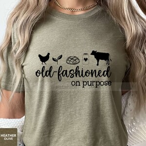 K&ouml;nnte beinhalten: Ein gr&uuml;nes T-Shirt mit dem Text "old-fashioned on purpose" und einer schwarzen Silhouette eines Huhns, einer Pflanze, eines Brotlaibs, eines Glases mit einem Herzen und einer Kuh.