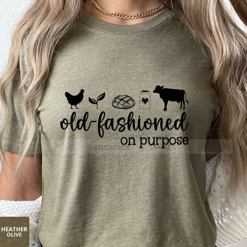 K&ouml;nnte beinhalten: Ein gr&uuml;nes T-Shirt mit dem Text "old-fashioned on purpose" und einer schwarzen Silhouette eines Huhns, einer Pflanze, eines Brotlaibs, eines Glases mit einem Herzen und einer Kuh.