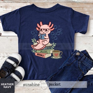 May include: A navy blue t-shirt with a pink axolotl wearing glasses and reading a book. The axolotl is sitting on a stack of books that say "I read a lotl books". There is a cup of coffee next to the books.