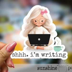 May include: A sticker featuring a cartoon illustration of a person with blonde hair, glasses, and a pink bow, sitting with a laptop. The sticker has the text "shhh. I'm writing" and a small teacup. The sticker is from "sunshine pocket".