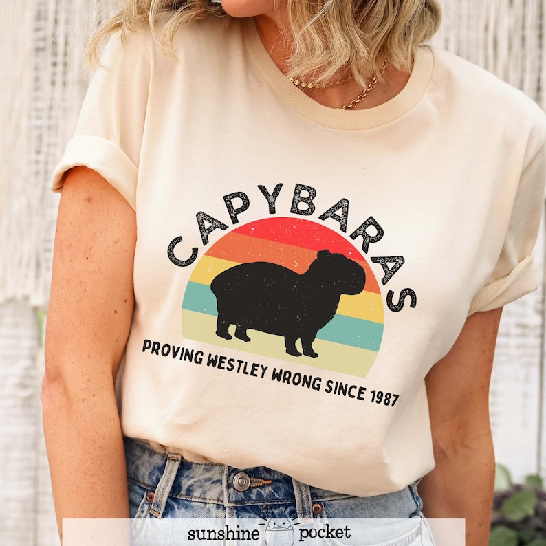 Pu&ograve; includere: Una maglietta beige con una grafica retr&ograve; di un capibara davanti a un tramonto. La grafica dice "Capybaras Proving Westley Wrong Since 1987".