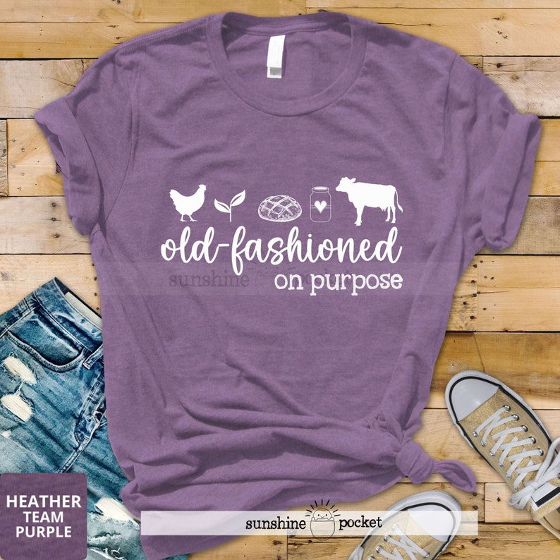 K&ouml;nnte beinhalten: Lilafarbenes T-Shirt mit einer wei&szlig;en Grafik einer Henne, einer Pflanze, einem Brotlaib, einem Glas mit einem Herzen und einer Kuh. Der Text lautet "old-fashioned sunshine on purpose".