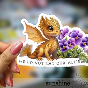 May include: A sticker featuring a cute, golden dragon with purple flowers. The dragon has large, expressive eyes and a friendly expression. The text on the sticker reads "We do not eat our allies."