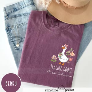 May include: Berry-coloured t-shirt featuring a goose graphic wearing glasses and holding books, with the text "Teacher Goose Mrs. Johnson." The shirt is styled with light blue jeans and a tan hat. A gold necklace is visible.