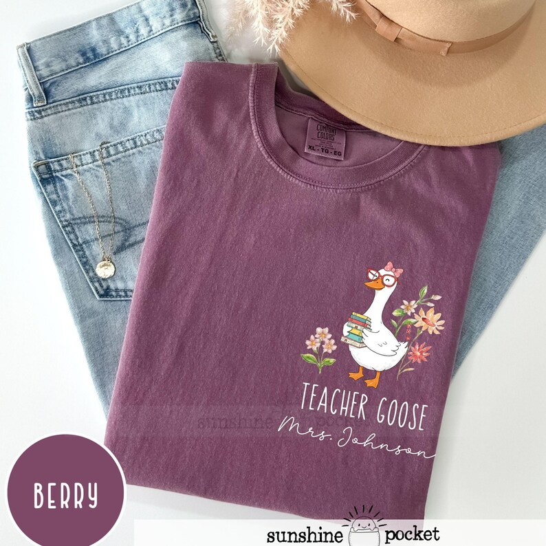 May include: Berry-coloured t-shirt featuring a goose graphic wearing glasses and holding books, with the text "Teacher Goose Mrs. Johnson." The shirt is styled with light blue jeans and a tan hat. A gold necklace is visible.