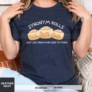 May include: A heather navy blue t-shirt featuring a graphic of three cartoon cinnamon rolls with white icing and smiling faces. The text above reads SYNONYM ROLLS and the text below reads JUST LIKE GRAMMAR USED TO MAKE in white lettering.