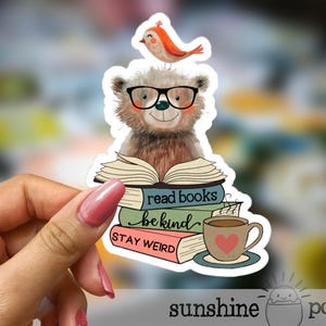 May include: A sticker featuring a cartoon bear wearing glasses with a bird perched on its head. The bear sits atop a stack of books with the phrases "read books", "be kind", and "stay weird". A cup of coffee with a heart is also shown.