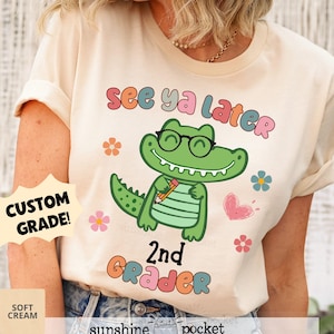 May include: A light beige t-shirt with the text "See ya later" in rainbow colors. A cartoon green alligator wearing glasses and holding a pencil is in the center of the shirt. The text "2nd Grader" is below the alligator. The shirt has a custom grade option.