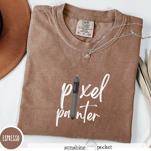 May include: Espresso-colored t-shirt with the words "pixel painter" and a graphic of a digital pen. The shirt has a crew neck and short sleeves. A silver necklace with a small pendant is draped across the shirt.