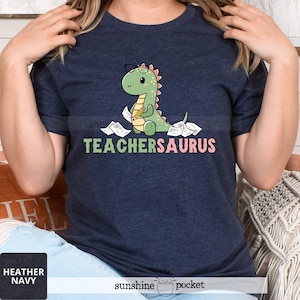May include: A navy blue t-shirt with a cartoon dinosaur wearing glasses and holding a pencil. The dinosaur is surrounded by papers. The text "TEACHERSAURUS" is printed in pink below the dinosaur.