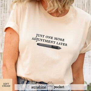 May include: Soft cream-colored t-shirt with the text "JUST ONE MORE ADJUSTMENT LAYER" and a graphic of a digital pen. The shirt also has the words "soft cream" and "sunshine pocket" printed on it.