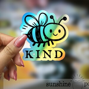 Peut inclure: Autocollant holographique avec une abeille de dessin animé souriante, aux contours noirs et aux couleurs bleu, vert et jaune. Le mot "KIND" est écrit en noir en dessous de l'abeille.