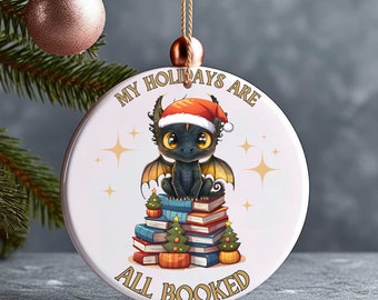 May include: Round Christmas ornament featuring a cartoon dragon wearing a Santa hat, sitting on a stack of books. The ornament has the text "MY HOLIDAYS ARE ALL BOOKED" and includes small Christmas trees. The ornament is hanging from a gold string.