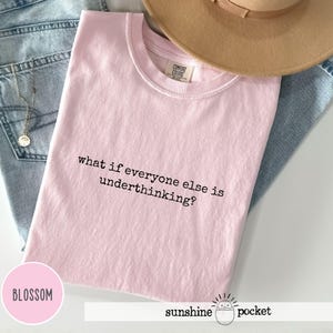 Peut inclure: Un t-shirt rose clair &agrave; manches courtes avec le texte imprim&eacute; en noir what if everyone else is underthinking? Le v&ecirc;tement est pr&eacute;sent&eacute; avec un tissu en jean bleu clair, un chapeau &agrave; larges bords beige et un collier &agrave; cha&icirc;ne dor&eacute;e.