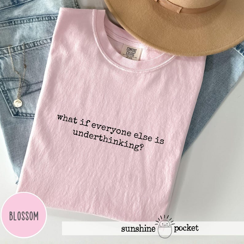 Peut inclure: Un t-shirt rose clair &agrave; manches courtes avec le texte imprim&eacute; en noir what if everyone else is underthinking? Le v&ecirc;tement est pr&eacute;sent&eacute; avec un tissu en jean bleu clair, un chapeau &agrave; larges bords beige et un collier &agrave; cha&icirc;ne dor&eacute;e.