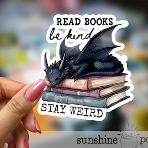 May include: A sticker featuring a black dragon resting on a stack of books. The sticker has the words "READ BOOKS be kind STAY WEIRD" in black text. The sticker is held by a hand with pink nail polish.