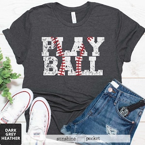 May include: A dark grey heather t-shirt with the text "PLAY BALL" in white with a red baseball graphic. The shirt is displayed on a white surface with a pair of white sneakers and blue denim shorts.