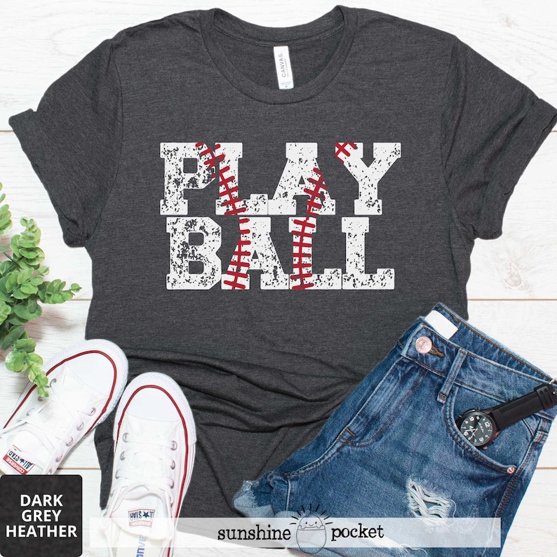 May include: A dark grey heather t-shirt with the text "PLAY BALL" in white with a red baseball graphic. The shirt is displayed on a white surface with a pair of white sneakers and blue denim shorts.