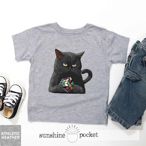 May include: Athletic heather gray t-shirt featuring a black cat holding a Rubik's Cube. The cat has yellow eyes and is depicted with a serious expression. The text "sunshine pocket" is printed at the bottom of the shirt.