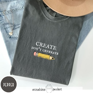 May include: A charcoal gray t-shirt featuring white embroidered text that reads CREATE DON'T GENERATE above a yellow pencil graphic with a pink eraser. The shirt is displayed alongside light blue denim jeans, a gold necklace, and a tan hat.