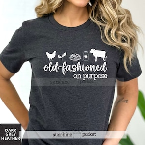 K&ouml;nnte beinhalten: Ein dunkel-graues T-Shirt mit einem wei&szlig;en Aufdruck eines Huhns, einer Pflanze, einem Brotlaib, einem Milchglas und einer Kuh. Unter dem Aufdruck steht der Text "old-fashioned on purpose".