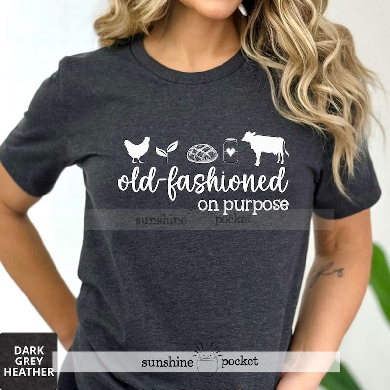 K&ouml;nnte beinhalten: Ein dunkel-graues T-Shirt mit einem wei&szlig;en Aufdruck eines Huhns, einer Pflanze, einem Brotlaib, einem Milchglas und einer Kuh. Unter dem Aufdruck steht der Text "old-fashioned on purpose".
