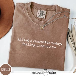 Peut inclure: T-shirt couleur expresso avec l'inscription blanche "killed a character today. feeling productive". Le t-shirt a un col rond et une petite étiquette. Un collier argenté avec un pendentif est drapé sur le t-shirt.